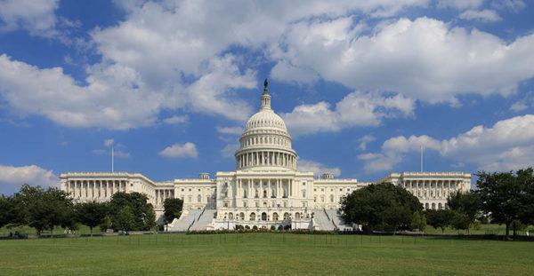 A photo of the United States Capitol under a cloudy blue sky and sitting on green grass. The capitol is white, with large stairs leading to the main floor. It has a large dome topping it.